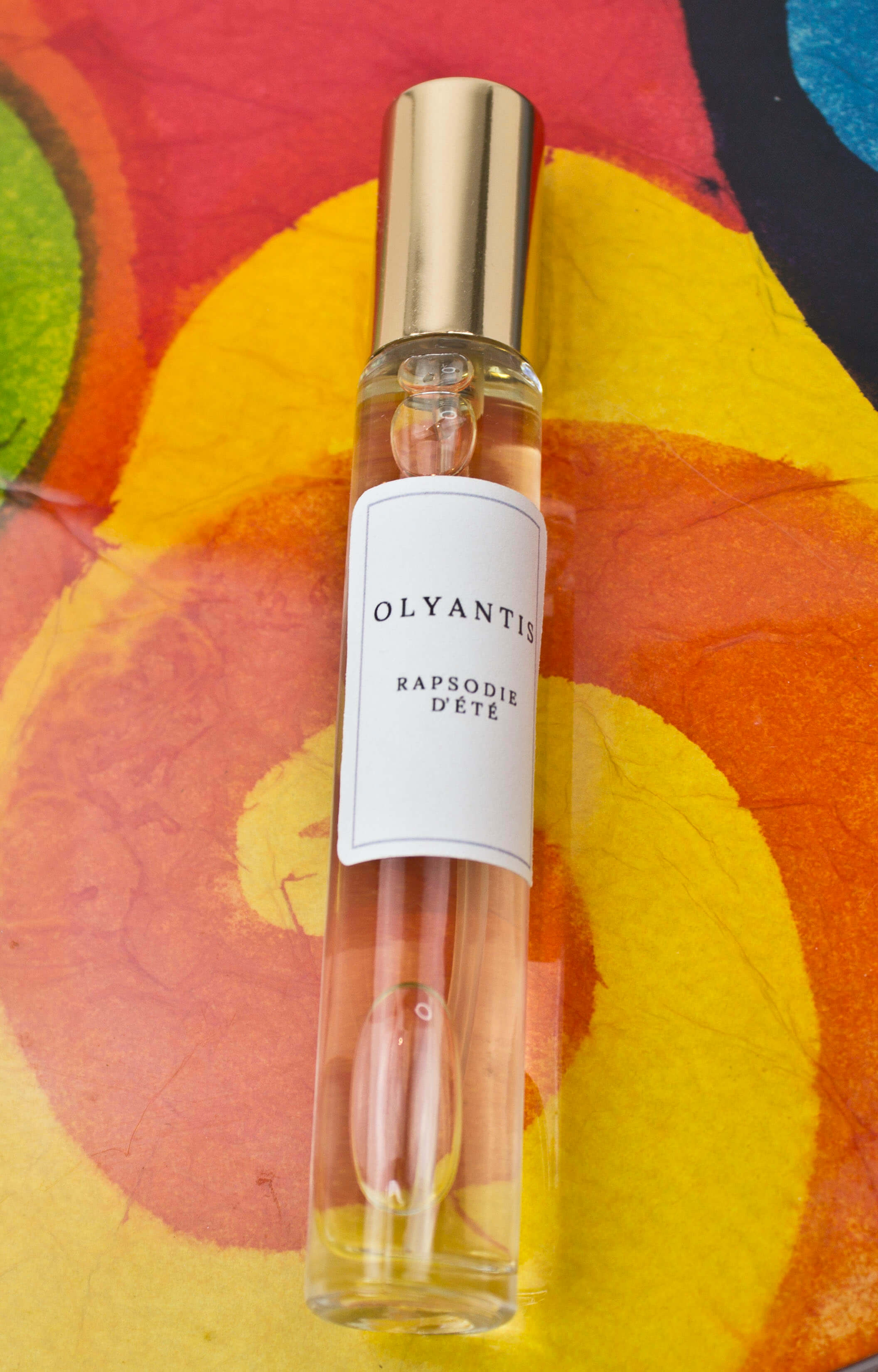 Olyantis's  Rapsodie D'Été 10 ml perfume glass bottle with gold cap on an orange, colorful background.
