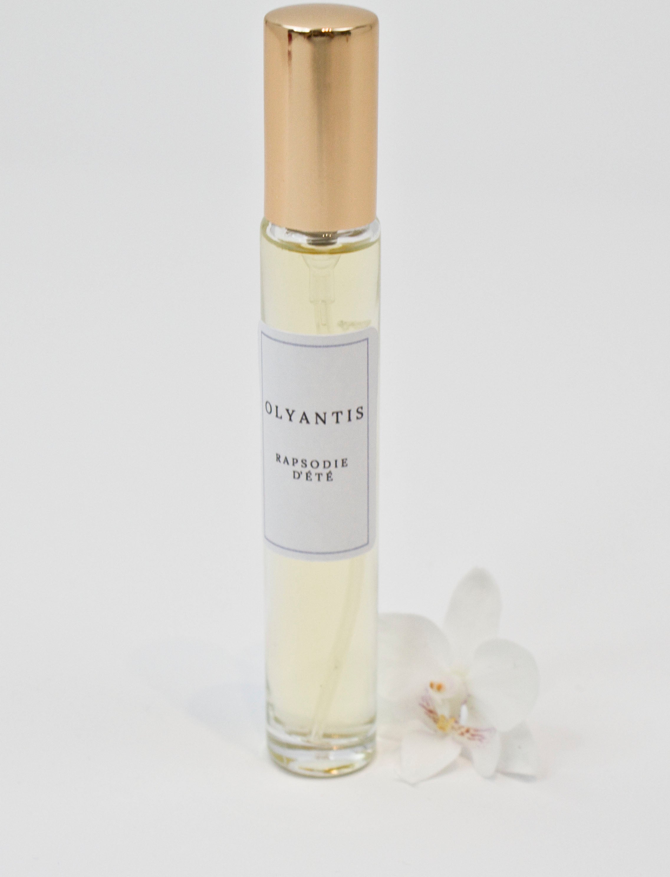 Olyantis's  Rapsodie D'Été 10 ml perfume glass bottle with gold cap beside a white flower.