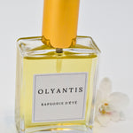 Olyantis's  Rapsodie D'Été 60 ml perfume glass bottle with gold cap beside a white flower.