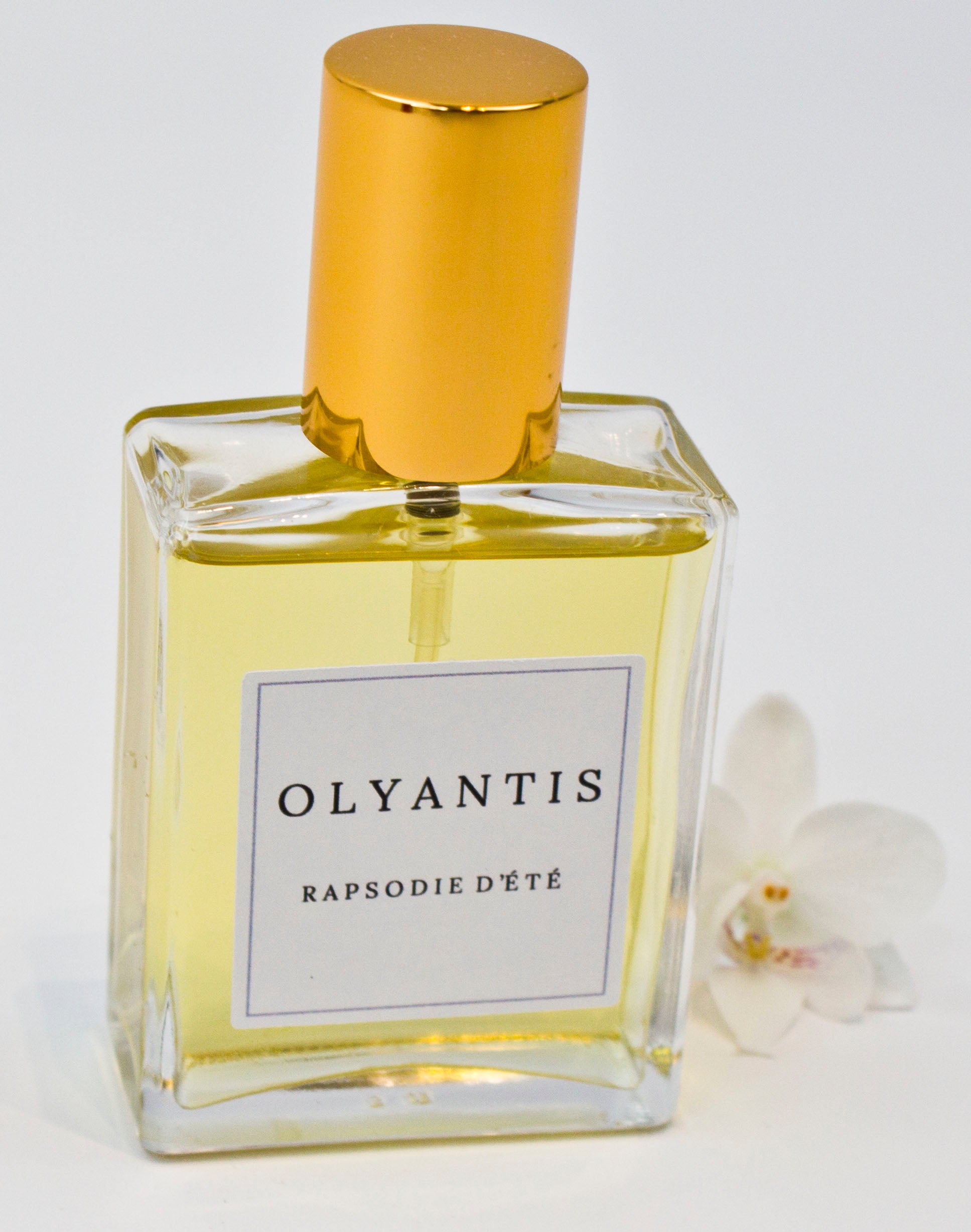 Olyantis's  Rapsodie D'Été 60 ml perfume glass bottle with gold cap beside a white flower.