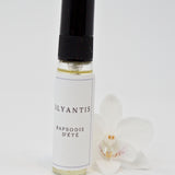 Olyantis's  Rapsodie D'Été 5 ml perfume glass bottle beside a white flower.
