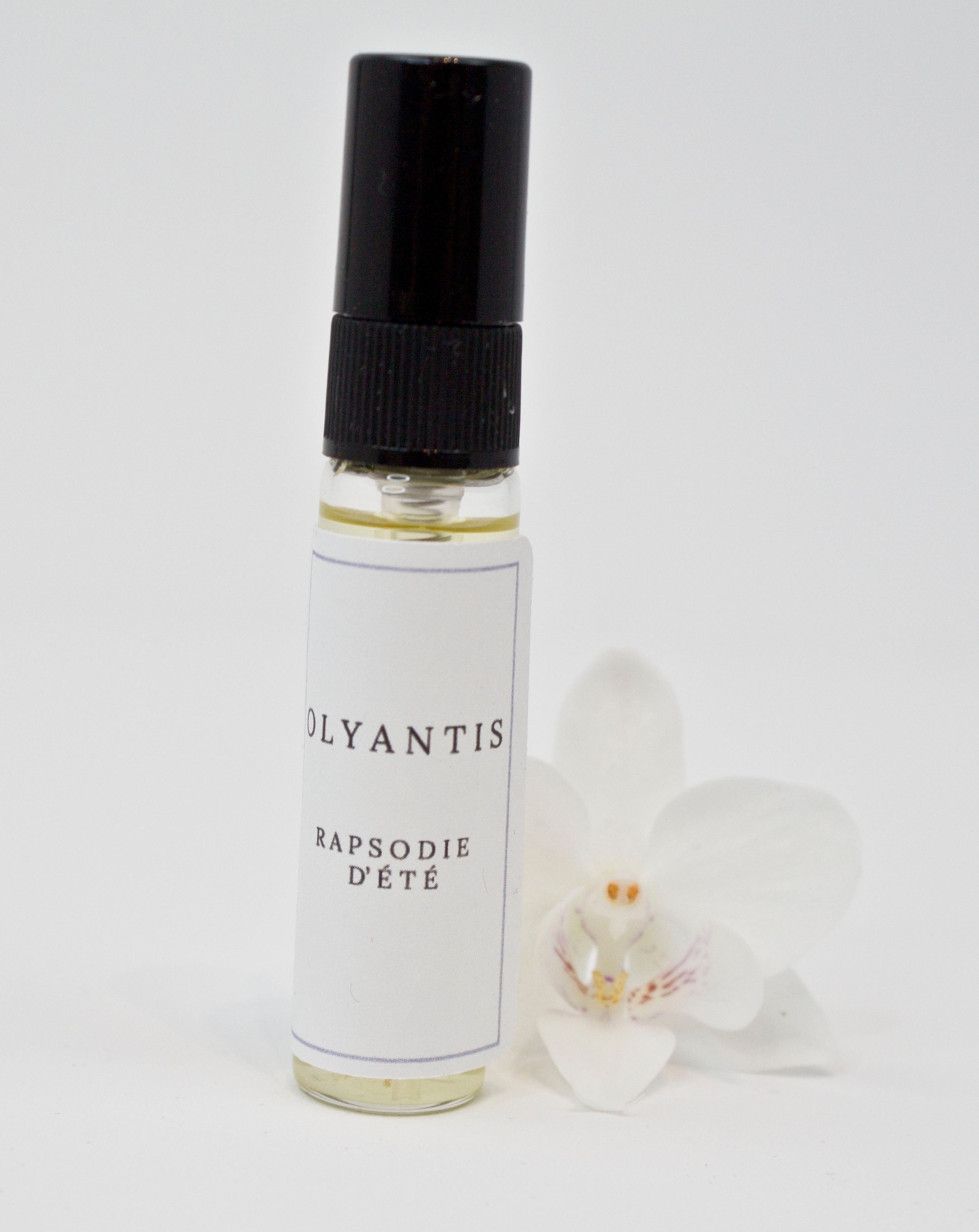Olyantis's  Rapsodie D'Été 5 ml perfume glass bottle beside a white flower.