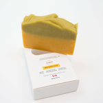 Rapsodie D'Été handmade, plant-based soap in a two-tone, sage and orange color, standing upright on top of its white cardboard packaging box against a clean white background.