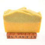 Rapsodie D'Été plant-based soap  bar standing upright on small wooden letter blocks that spell 'OLYANTIS'. The soap has a two-tone color identity with a gentle sage top layer and a warm orange-yellow bottom layer.