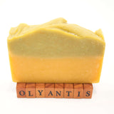 Rapsodie D'Été plant-based soap  bar standing upright on small wooden letter blocks that spell 'OLYANTIS'. The soap has a two-tone color identity with a gentle sage top layer and a warm orange-yellow bottom layer.