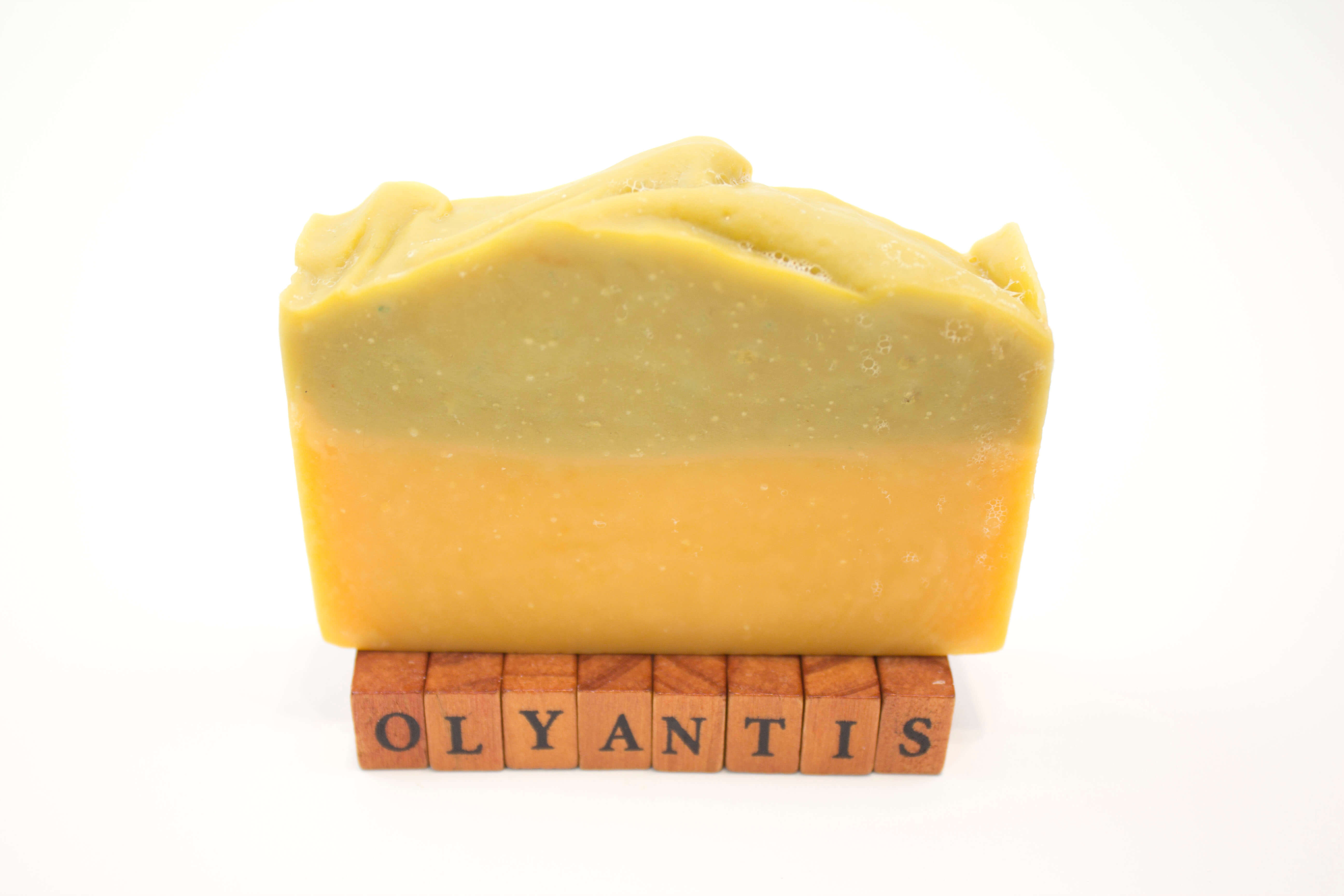 Rapsodie D'Été plant-based soap  bar standing upright on small wooden letter blocks that spell 'OLYANTIS'. The soap has a two-tone color identity with a gentle sage top layer and a warm orange-yellow bottom layer.