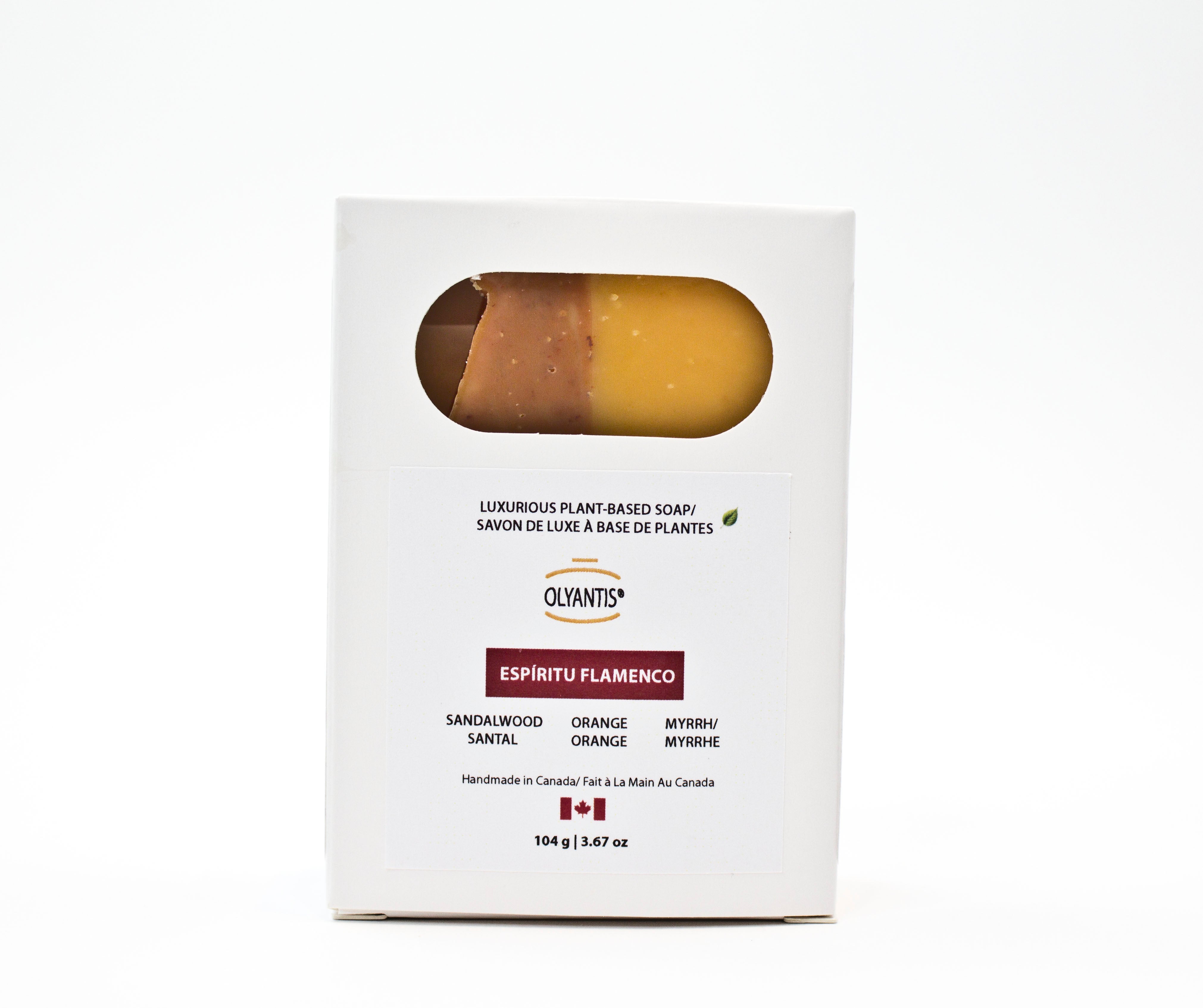 A white rectangular box of Olyantis's Espiritu Flamenco luxurious plant-based soap, featuring a clear oval window that shows the amber & golden-yelllow soap bar inside. The box also shows scent notes of sandalwood, myrrh and orange, and a small Canadian flag near the bottom.